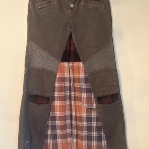 May include: A unique, olive-green denim skirt with a button closure and a plaid fabric panel. The skirt features a patchwork design with textured panels and pockets, creating a stylish, layered look. The skirt has a zipper on the side.