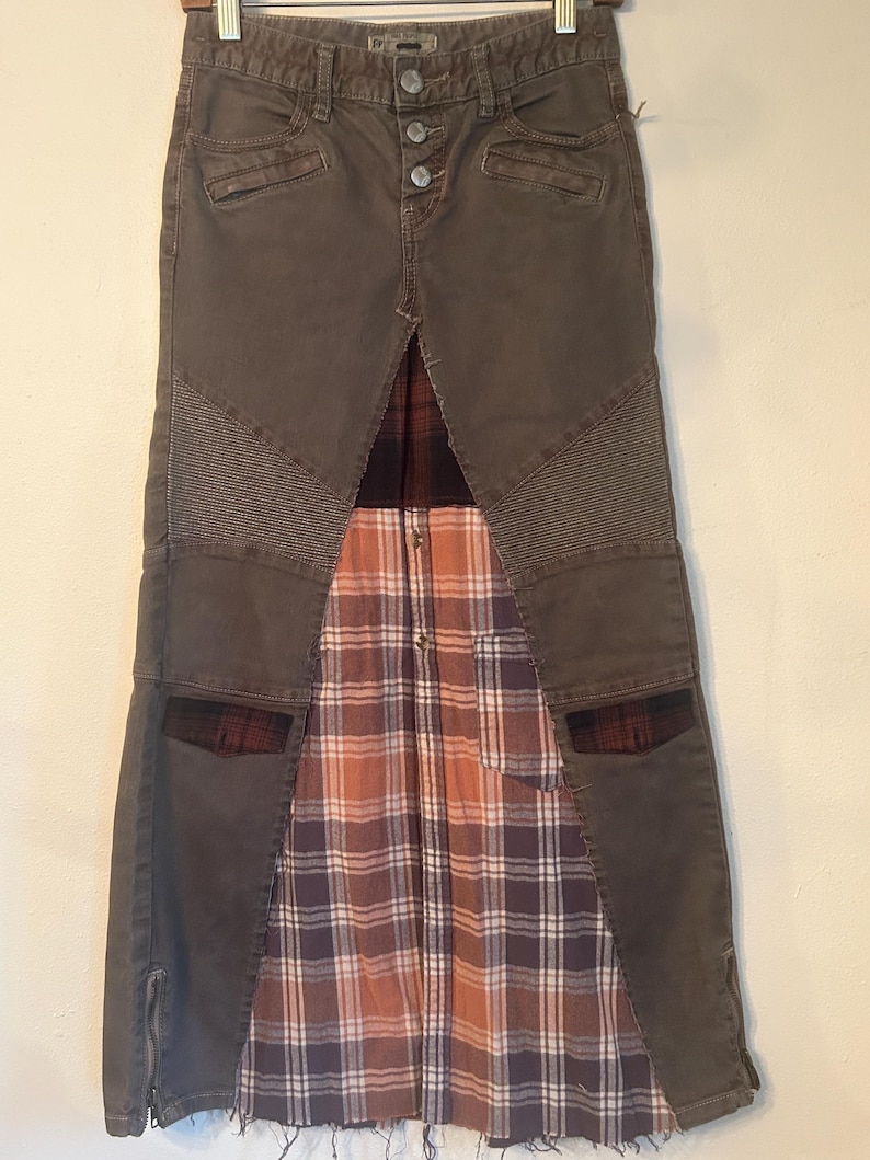 May include: A unique, olive-green denim skirt with a button closure and a plaid fabric panel. The skirt features a patchwork design with textured panels and pockets, creating a stylish, layered look. The skirt has a zipper on the side.