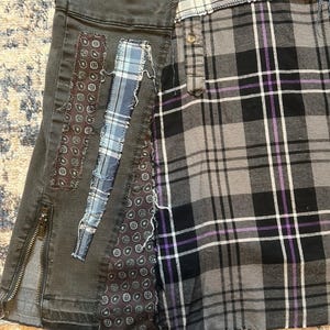 Puede incluir: Una falda patchwork que combina tejidos vaqueros y de cuadros. La secci&oacute;n de mezclilla gris oscuro tiene una cremallera lateral y parches decorativos. La secci&oacute;n de cuadros tiene un patr&oacute;n de cuadros gris, negro y morado.