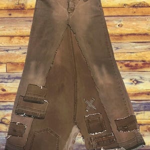 May include: Brown denim skirt with a unique patchwork design. The skirt features a high waist, a long length, and multiple pockets. The patchwork includes various denim pieces with frayed edges and decorative stitching.
