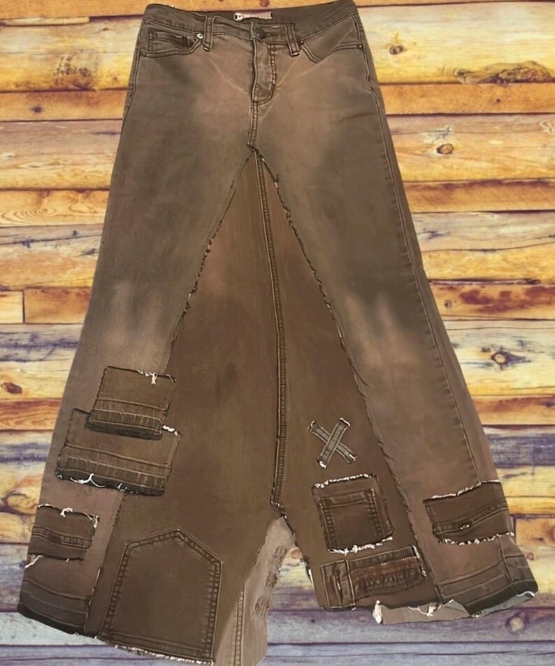 May include: Brown denim skirt with a unique patchwork design. The skirt features a high waist, a long length, and multiple pockets. The patchwork includes various denim pieces with frayed edges and decorative stitching.