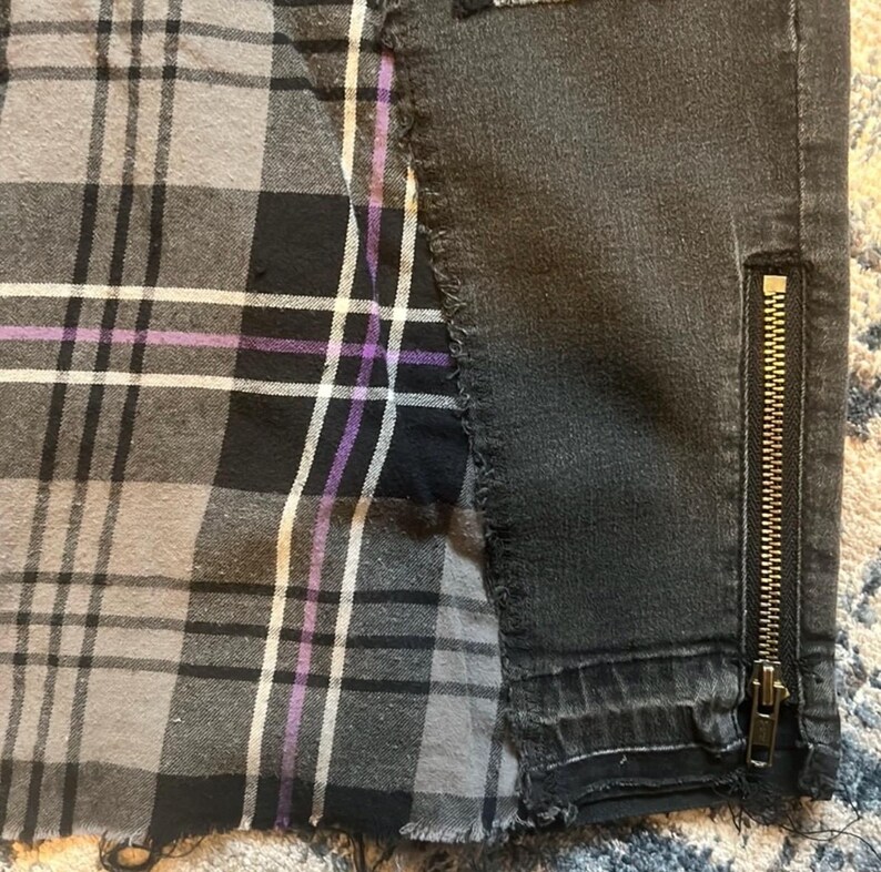 May include: A pair of black denim pants with a plaid panel. The plaid panel features a gray, black, white, and purple pattern. The pants have a zipper detail on the side.