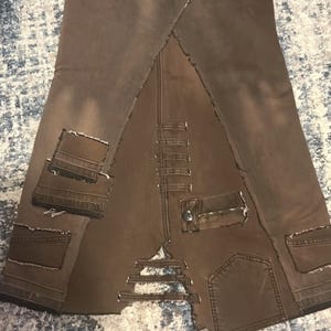 May include: Brown denim skirt with a unique patchwork design. Features multiple pockets, distressed edges, and a zipper detail. The skirt has a distinctive, deconstructed aesthetic.