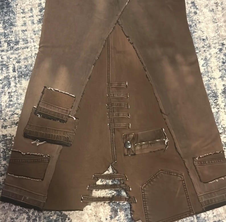 May include: Brown denim skirt with a unique patchwork design. Features multiple pockets, distressed edges, and a zipper detail. The skirt has a distinctive, deconstructed aesthetic.