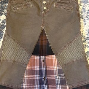 May include: Brown denim pants with a plaid shirt panel. The pants have button closures, pockets, and quilted accents on the knees. The plaid shirt features a button-down design with shades of brown, red, and blue.