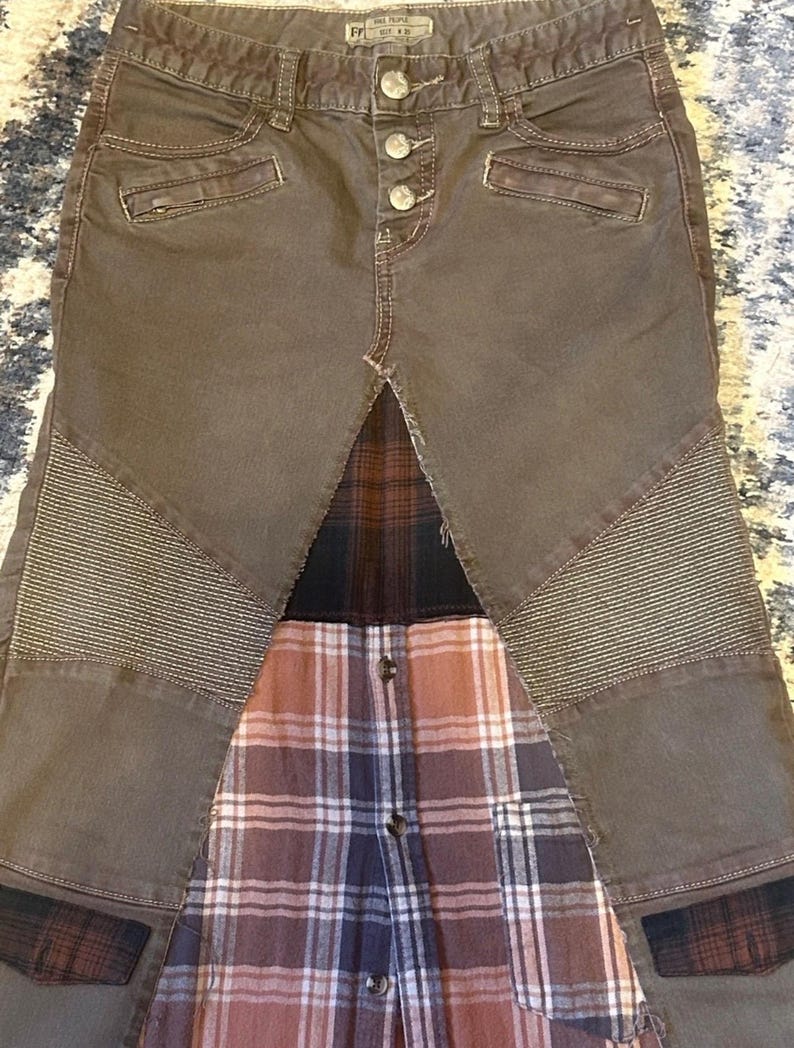 May include: Brown denim pants with a plaid shirt panel. The pants have button closures, pockets, and quilted accents on the knees. The plaid shirt features a button-down design with shades of brown, red, and blue.