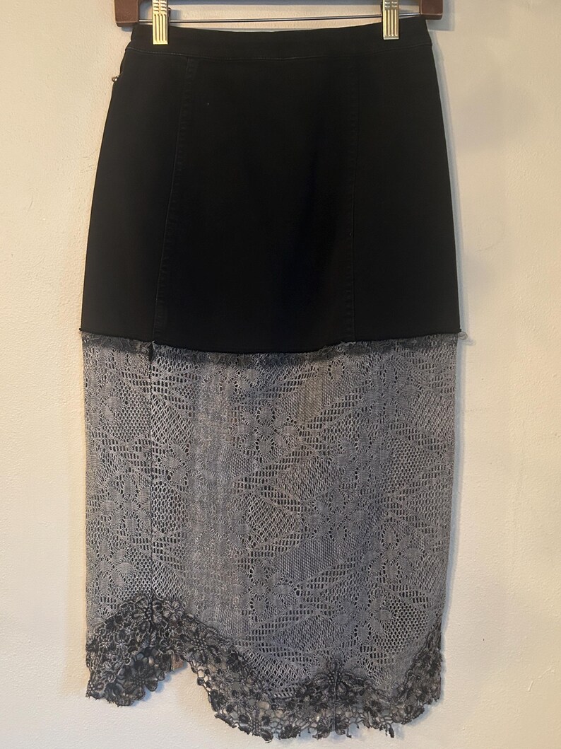 K&ouml;nnte beinhalten: Schwarzer Jeansrock mit grauem Spitzen-Overlay. Der Rock h&auml;ngt an einem Holzb&uuml;gel. Das Spitzen-Overlay hat ein Blumenmuster und einen gewellten Saum. Der Rock ist im Bleistiftrock-Stil.