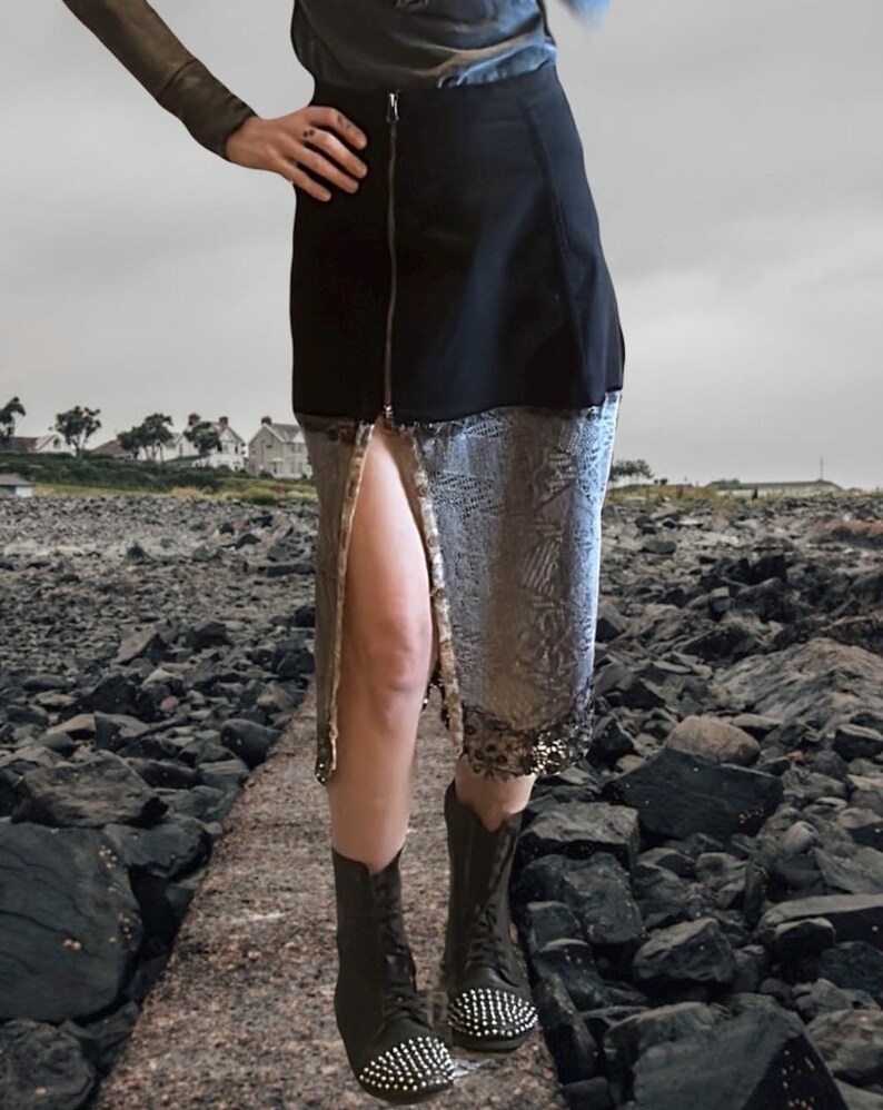 K&ouml;nnte beinhalten: Schwarzer Rock mit silbernem Paillettenbesatz und Frontrei&szlig;verschluss. Der Rock wird mit schwarzen Stiefeletten mit silbernen Nieten getragen. Das Outfit befindet sich vor einem felsigen Hintergrund im Freien.