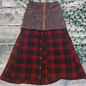 May include: A two-tone skirt featuring a leopard print denim top with a gold zipper and a red and black plaid bottom. The skirt has a button-down front and a unique, layered design.