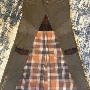 May include: A unique skirt design featuring a combination of materials. The top portion is a dark gray denim with button and zipper details. The skirt's lower section incorporates a plaid pattern in shades of brown, white, and gray.