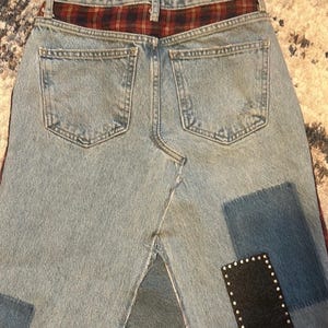 May include: Light blue denim skirt with a red and black plaid waistband. The skirt features two back pockets and decorative patches in dark blue and black with silver studs. The skirt has a split in the back.