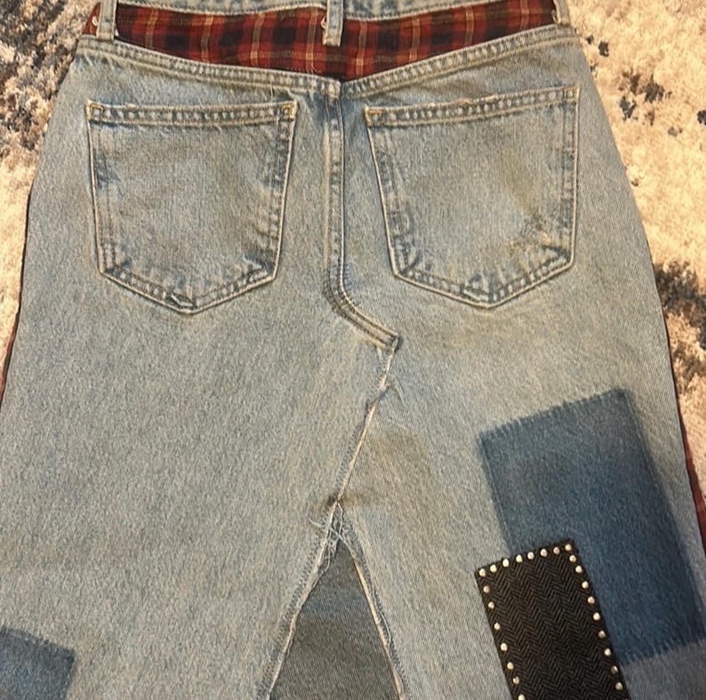 May include: Light blue denim skirt with a red and black plaid waistband. The skirt features two back pockets and decorative patches in dark blue and black with silver studs. The skirt has a split in the back.
