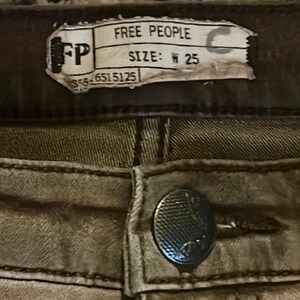 May include: Close-up of a pair of olive-green denim pants. The tag reads "FREE PEOPLE" and "SIZE: W 25". A silver button is visible below the tag.