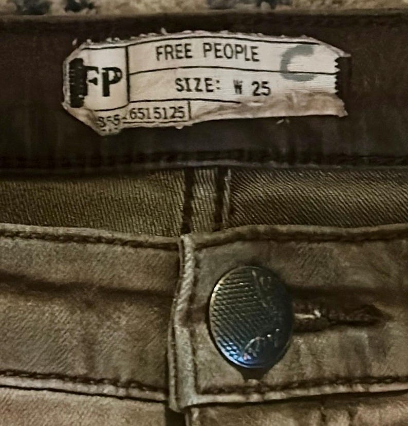 May include: Close-up of a pair of olive-green denim pants. The tag reads "FREE PEOPLE" and "SIZE: W 25". A silver button is visible below the tag.