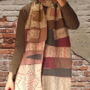 Puede incluir: Bufanda larga de patchwork en tonos beige, marrón y burdeos. La bufanda presenta una variedad de texturas y patrones de tela, incluyendo cuadrados, rectángulos y formas de diamante. La bufanda es larga y parece hecha a mano.