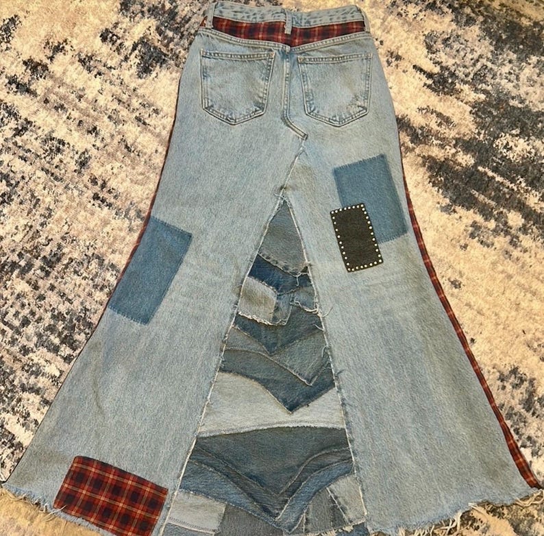 May include: A light blue denim skirt with a patchwork design. The skirt features a high waist with a red and black plaid waistband. Patches of denim and a black square with white dots are sewn onto the skirt.