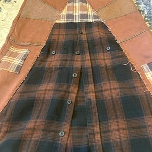 May include: A unique, handmade skirt featuring a patchwork design. The skirt combines brown denim panels with a black and brown plaid flannel shirt. Additional plaid patches and textured fabric add detail. The skirt has a button-down front.