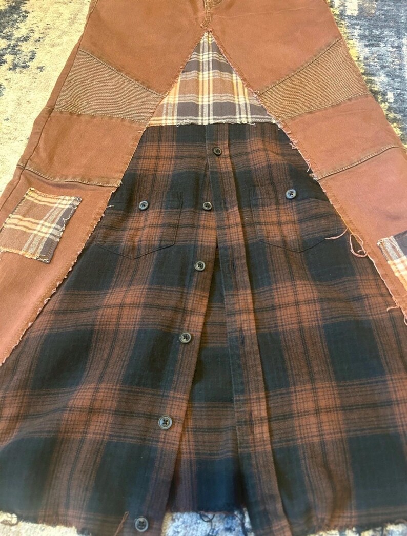 May include: A unique, handmade skirt featuring a patchwork design. The skirt combines brown denim panels with a black and brown plaid flannel shirt. Additional plaid patches and textured fabric add detail. The skirt has a button-down front.