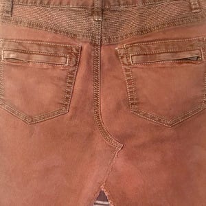 May include: Close-up of the back of a rust-colored denim skirt. Features include two back pockets with zippers, decorative stitching, and a split hem revealing a plaid fabric insert. The skirt is hanging on a hanger.