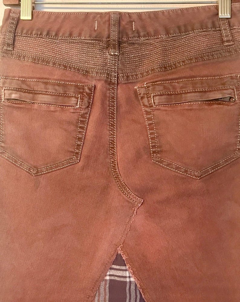 May include: Close-up of the back of a rust-colored denim skirt. Features include two back pockets with zippers, decorative stitching, and a split hem revealing a plaid fabric insert. The skirt is hanging on a hanger.