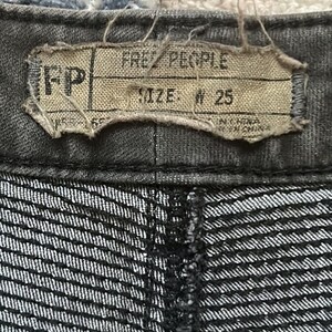Puede incluir: Primer plano de unos pantalones vaqueros grises oscuros. La etiqueta dice "FREE PEOPLE" y "SIZE: W 25". La tela tiene un sutil estampado de rayas.