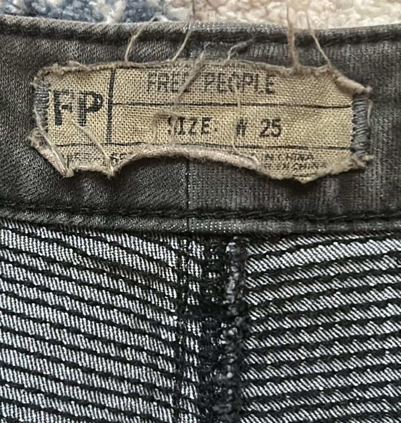 Puede incluir: Primer plano de unos pantalones vaqueros grises oscuros. La etiqueta dice "FREE PEOPLE" y "SIZE: W 25". La tela tiene un sutil estampado de rayas.