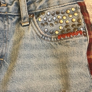 May include: Close-up of light-wash denim jeans with a studded pocket. The pocket is embellished with numerous silver and gold-toned studs. Red stitching accents the pocket's edge, and a plaid fabric detail is visible on the side.