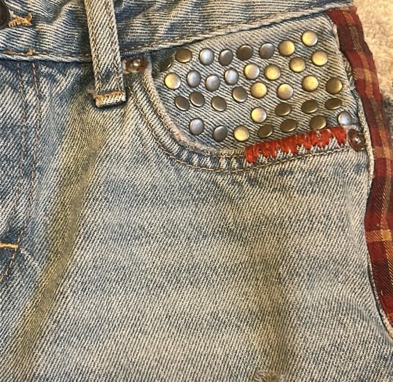 May include: Close-up of light-wash denim jeans with a studded pocket. The pocket is embellished with numerous silver and gold-toned studs. Red stitching accents the pocket's edge, and a plaid fabric detail is visible on the side.