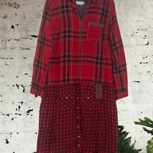 Könnte beinhalten: Ein langärmliges rotes kariertes Kleid mit Knopfleiste und zwei Vordertaschen. Das Kleid hat ein Patchwork-Design mit einer Mischung aus roten und schwarzen Karomustern. Das Kleid hängt an einem Holzkleiderbügel.