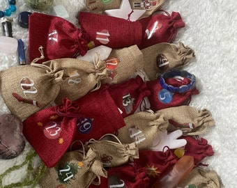 Op de afbeelding: Een jute adventskalender met rode en bruine stoffen zakjes. Elk zakje heeft een nummer van 1 tot 24 erop gedrukt. De kalender is omringd door verschillende kleurrijke stenen en kristallen.