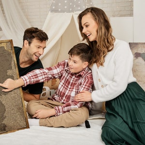 May include: A family of three is looking at a vintage-style map of the Netherlands. The map is framed and hanging on a wall. The family is sitting on a white bed with a white canopy. The map is a scratch-off map, and the family is scratching off the places they have visited.