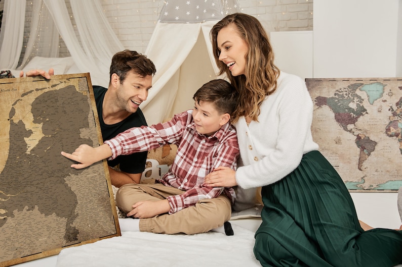 May include: A family of three is looking at a vintage-style map of the Netherlands. The map is framed and hanging on a wall. The family is sitting on a white bed with a white canopy. The map is a scratch-off map, and the family is scratching off the places they have visited.