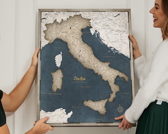 May include: A framed map of Italy with a blue background and beige lines showing the country's regions. The map is titled "Italia" in gold lettering. Two people are holding the map up against a white wall.