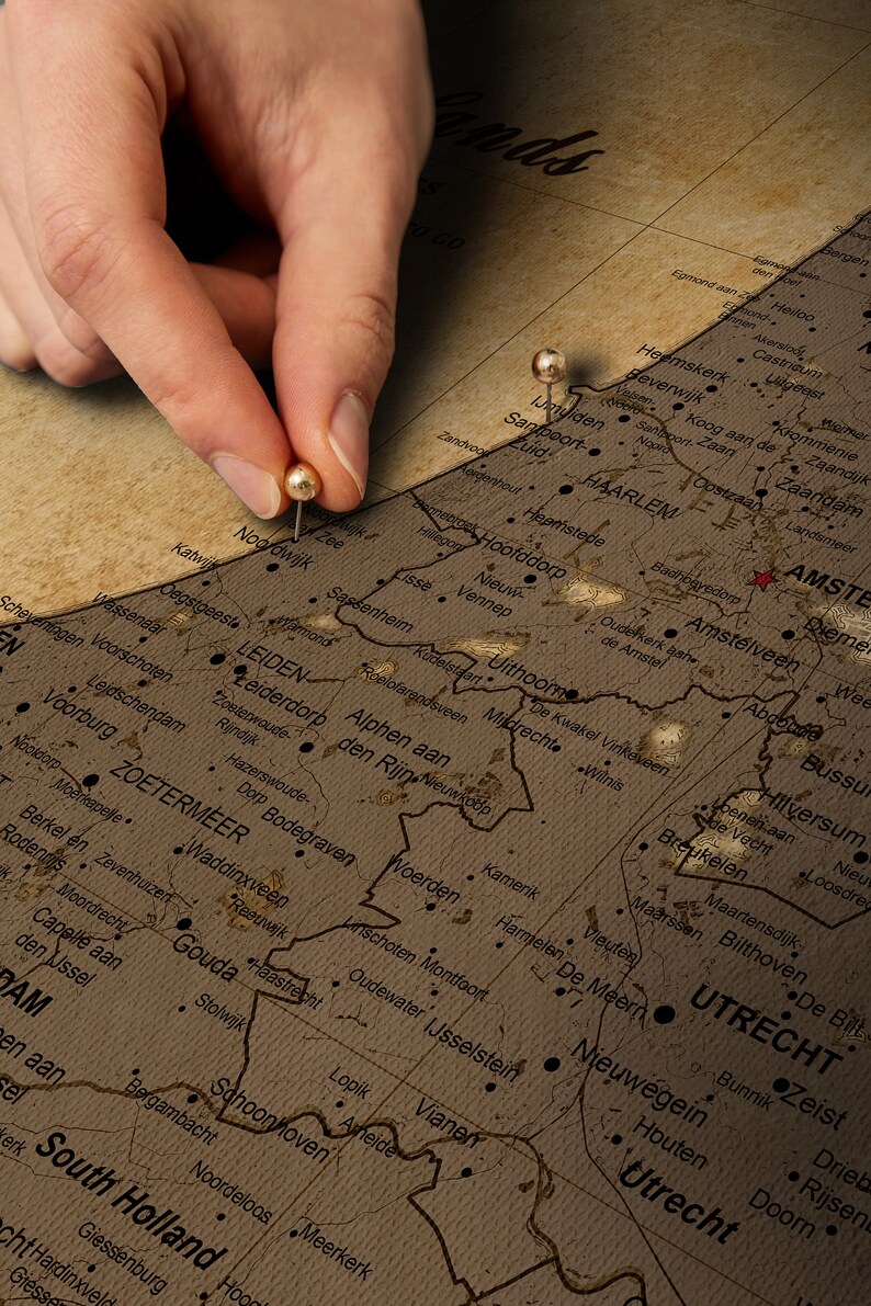 May include: A close-up of a vintage map of the Netherlands with push pins marking the cities of Leiden and Amsterdam. The map is on a brown background and has a worn, aged look.