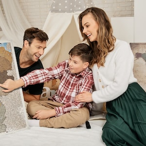 May include: A family of three is looking at a map of Europe. The map is on a white surface and the family is sitting on a white rug. The map is a scratch-off map, and the family has scratched off some of the countries. The map is a great way to track your travels.