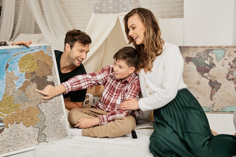 May include: A family of three is looking at a map of Europe. The map is on a white surface and the family is sitting on a white rug. The map is a scratch-off map, and the family has scratched off some of the countries. The map is a great way to track your travels.