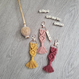 May include: Three colorful macrame keychains with gold key rings and a tassel at the bottom. The keychain on the left is yellow, the middle keychain is pink, and the keychain on the right is red. The pink keychain has a wooden bead with the name "Lucy" spelled out on it. The wooden beads above the keychains spell out "Dein Wunsch Name".