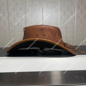 May include: A brown leather cowboy hat with a wide brim. The hat has three small air vents on the crown and a decorative silver accent. A leather tie is attached to the brim. The hat is displayed on a black surface.