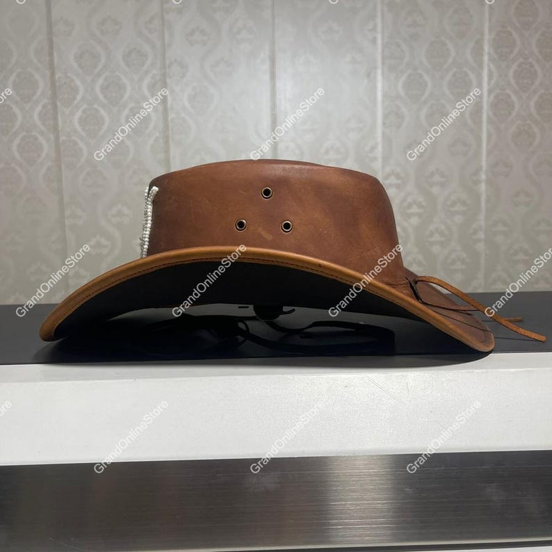May include: A brown leather cowboy hat with a wide brim. The hat has three small air vents on the crown and a decorative silver accent. A leather tie is attached to the brim. The hat is displayed on a black surface.