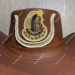 May include: A brown leather cowboy hat with an embroidered Egyptian design. The design features a gold and pearl-like border, with an Egyptian figure at the centre. The hat is a classic Western style.