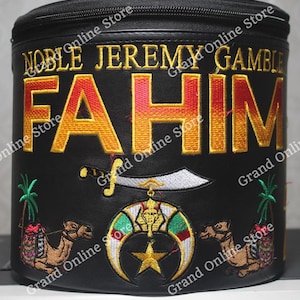 Puede incluir: Fez de cuero negro con bordados dorados. El fez tiene el nombre "NOBLE JEREMY FAHIM GAMBLE" bordado en él. El fez también tiene una luna creciente y una estrella, una espada y un camello bordados.