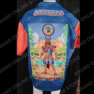 May include: Blue button-up shirt with orange sleeves and a large graphic on the back. The graphic features an illustration of an Anubis-like figure, pyramids, and palm trees. The shirt has the word "LEGENDS" across the top.