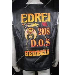 以下が含まれることがあります： 刺繍されたテキストとシンボルが付いた黒い革のベスト。「EDREI」の文字が黄色で、背面に「NO. 2108 D.O.S. GEORGIA」と表示されています。 剣と三日月のデザインも表示されています。