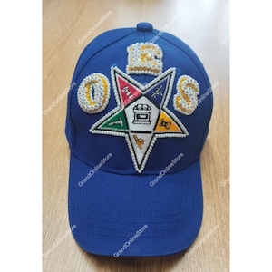 Puede incluir: Una gorra de béisbol azul con visera curva. La gorra presenta símbolos bordados y las letras "OES" en blanco. Un gran emblema en forma de estrella está centrado en la parte delantera, con varios símbolos y un borde de cuentas blancas. El texto "GrandOnlineStore" está impreso.