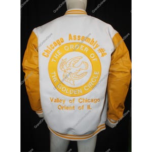 Op de afbeelding: Witte en gouden varsity jas met de tekst "Chicago Assembly #4 The Order of the Golden Circle" geborduurd op de rug. De jas heeft gouden mouwen en afwerking.