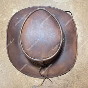 May include: A brown leather cowboy hat with a rounded crown and a wide brim. The hat has a distressed appearance and a chin strap. The text "GrandOnlineStore" is visible on the hat.