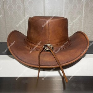 May include: A brown leather cowboy hat with a wide brim and a tall crown. The hat has a decorative silver-coloured buckle and a leather chin strap. The hat is a rich, warm brown colour.