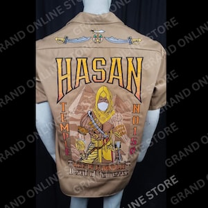 May include: Tan short-sleeve shirt with embroidered design. The back features the name "HASAN" above an illustration of a figure with a pyramid background. Additional text includes "TEMPLE" and "N0157".