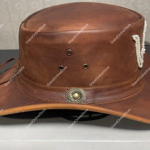 May include: A brown leather hat with a wide brim and a decorative band around the crown. The hat features a metal emblem and a string of white beads. The hat has three small eyelets and is designed for sun protection.