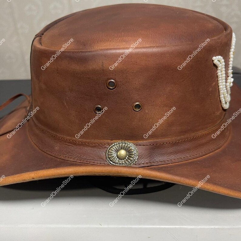 May include: A brown leather hat with a wide brim and a decorative band around the crown. The hat features a metal emblem and a string of white beads. The hat has three small eyelets and is designed for sun protection.